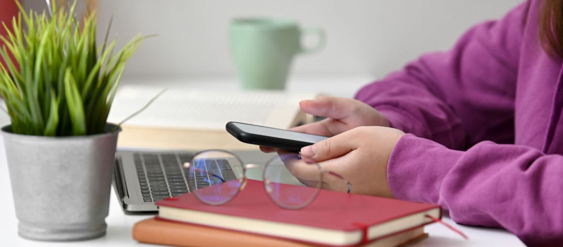 Side view of college student relaxing with mock-up smartphone while doing assignment with supplies and decoration on white table