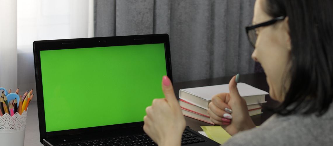 Woman teacher looking at web camera making video call on laptop with green screen, chroma key. Working online from home. Distance education with children class on internet. Conference training webinar