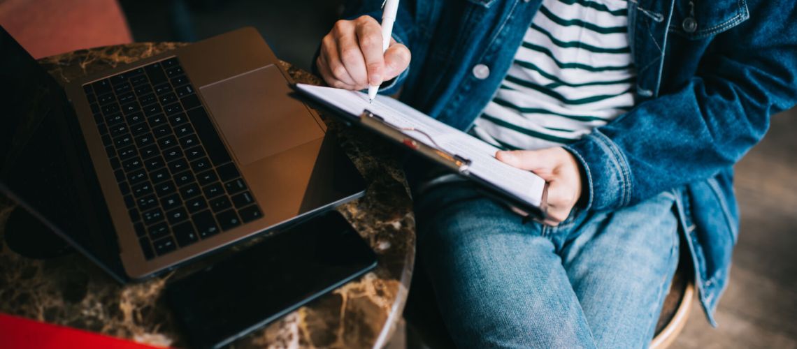 From above crop modern man in casual outfit sitting with crossed legs at table and taking notes in clipboard working with with laptop in cafe