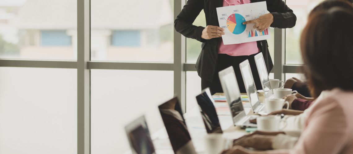 Group of businesswomen meeting together in office, leader or manager holding graph and chart while explaining details and meaning to audiences, the colleagues listen to her and express understanding.